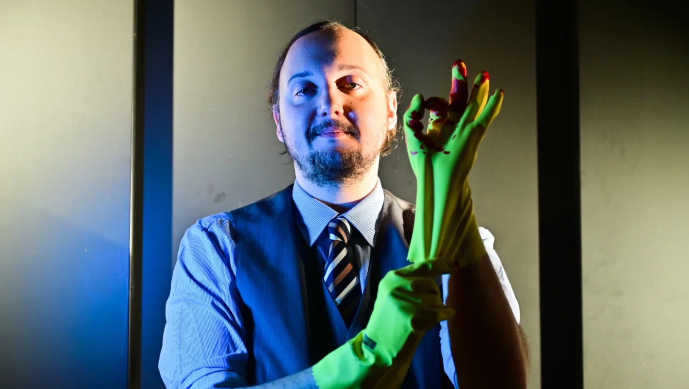 Julian Niedermeier in edler Kleidung zieht sich einen blutverschmierten gelben Reinigungshandschuh an