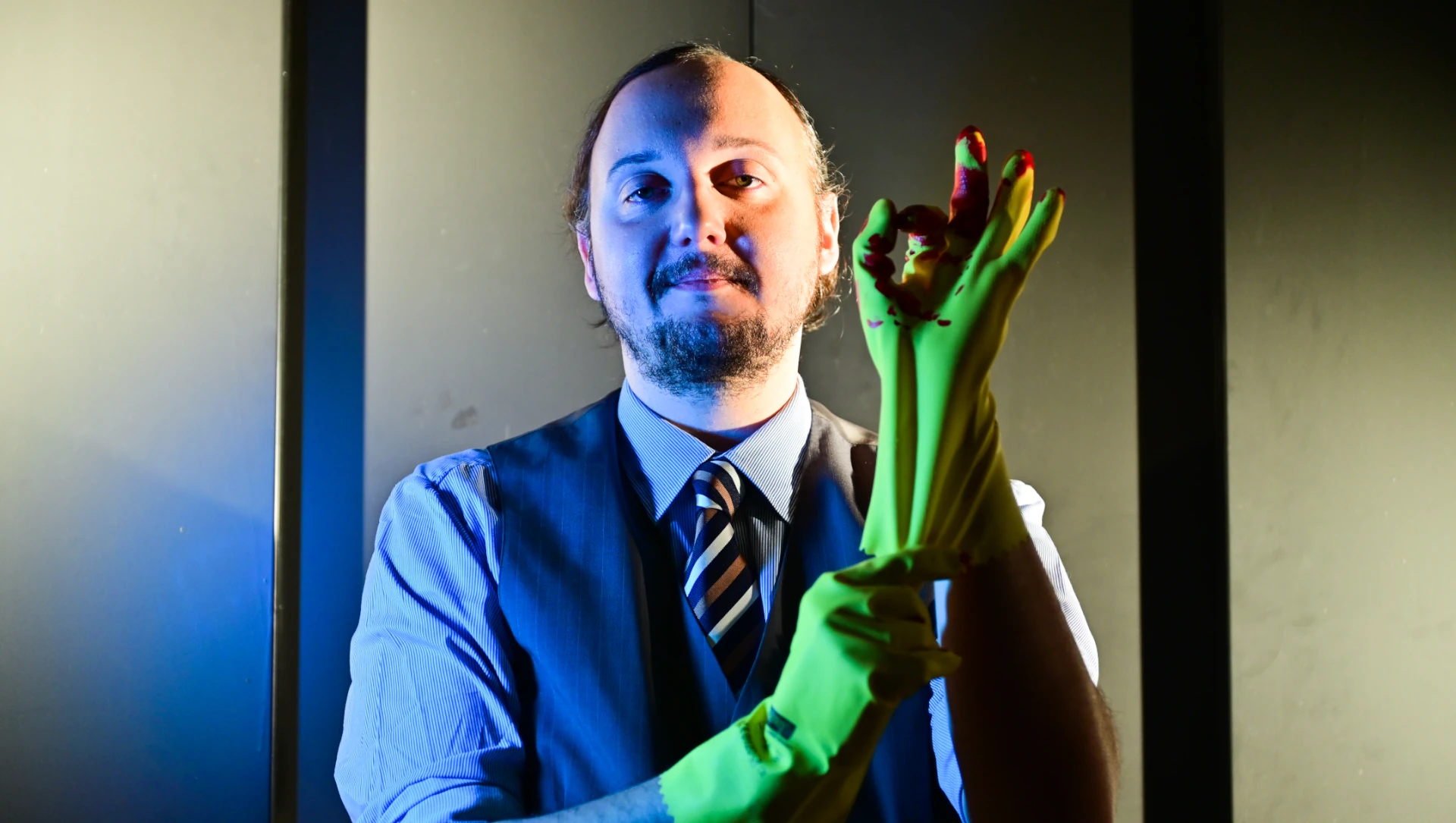 Julian Niedermeier in edler Kleidung zieht sich einen blutverschmierten gelben Reinigungshandschuh an