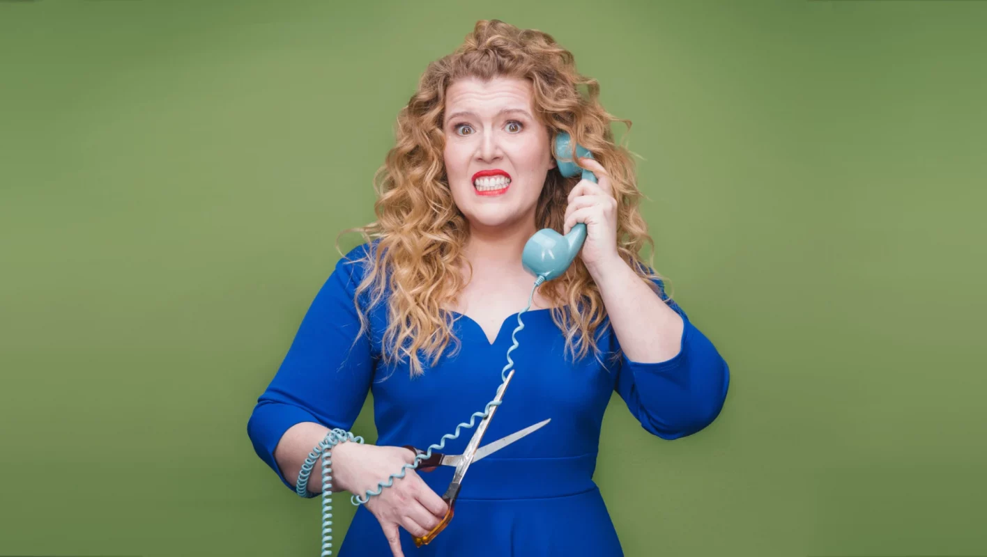 Franziska Wanninger im blauen kleit hält sich rechts einen Telefonhörer ans Ohr. In der linken Hand hält sie eine Schere um das Telefonkabel durchzuschneiden.