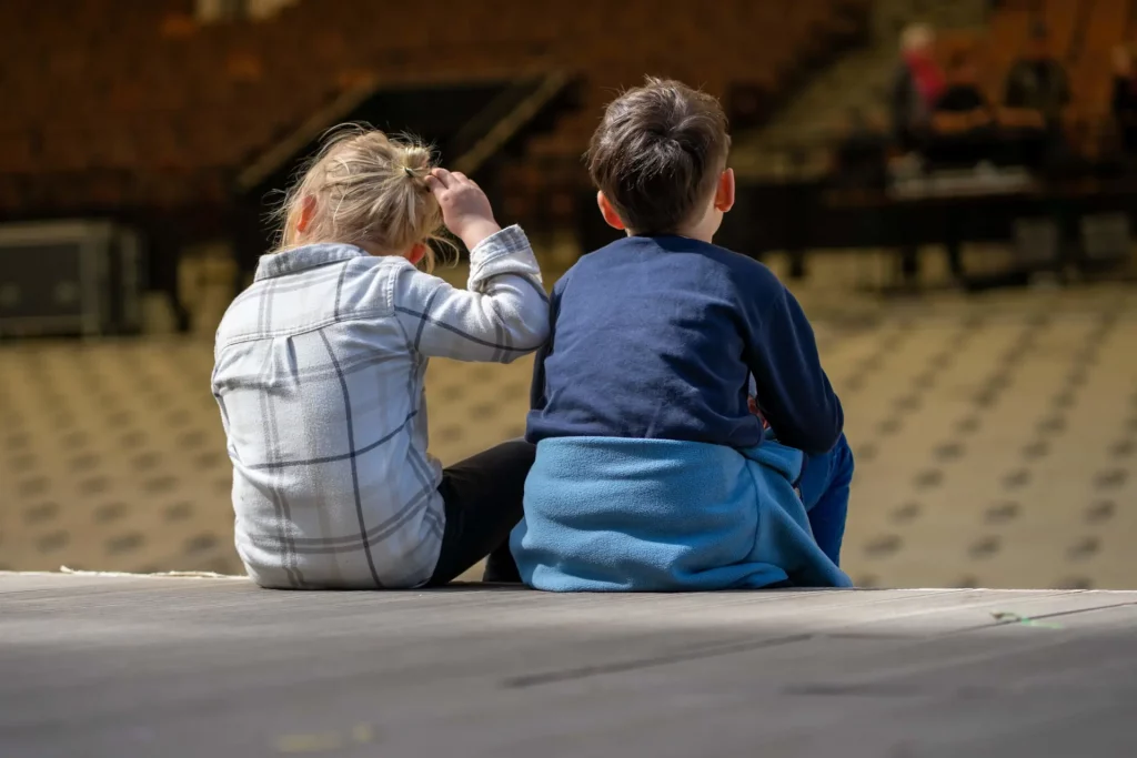 Blick von hinten auf zwei Kinder die auf der Bühnenkante sitzen. Im Hintergrund der Zuschauerraum.
