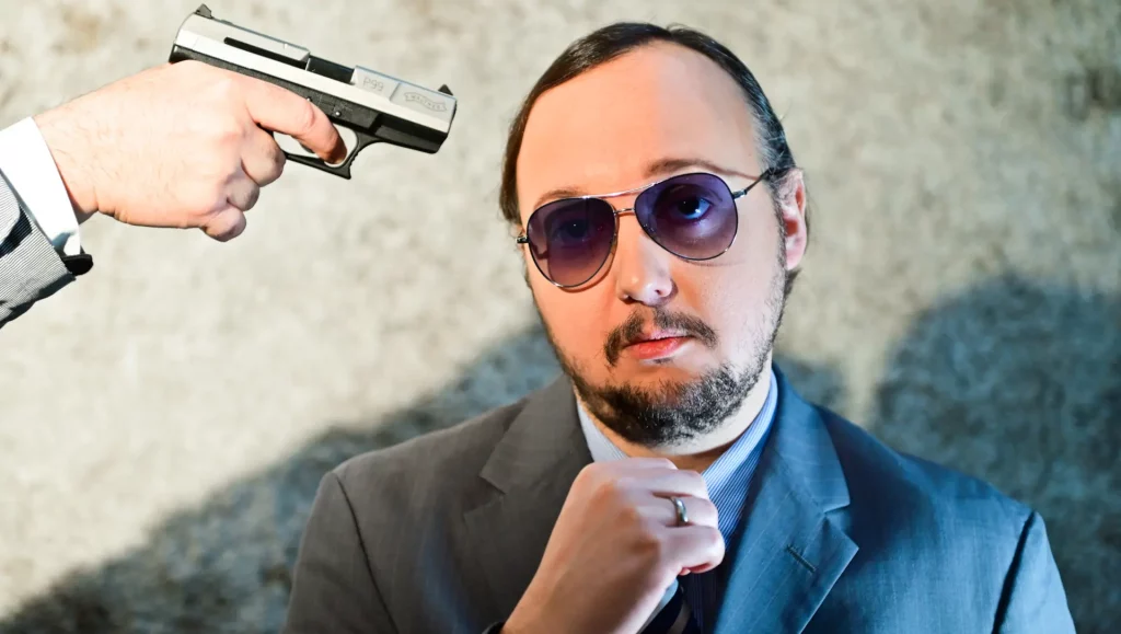 Ein Mann im Anzug mit Sonnenbrille blickt gelassen in die Kamera, während von Links oben eine Pistole auf seinen Kopf zielt.
